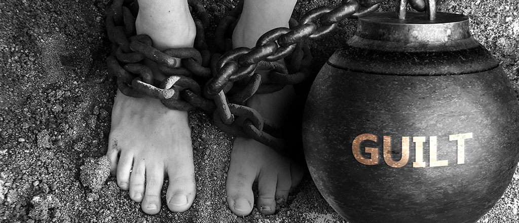Füße einer Person, die eine schwere Kette mit einer Kugel trägt, auf der "guilt" steht.